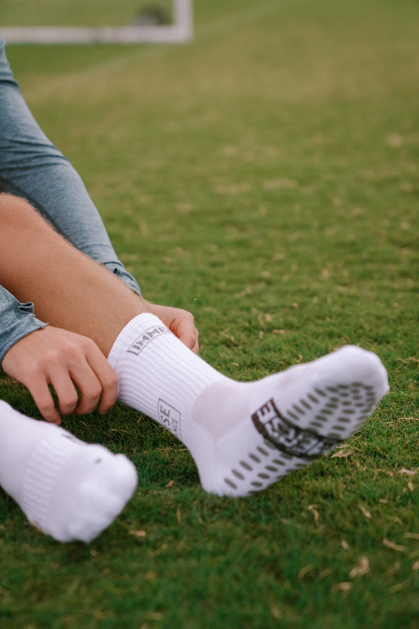 A player pulling on a white Immerse grip crew sock on the grass, the grip pads on the sole facing the camera