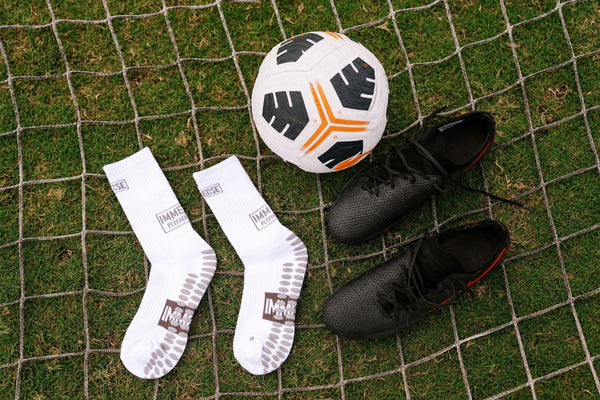 White Immerse GRIP crew socks on a goal net beside a ball and black boots